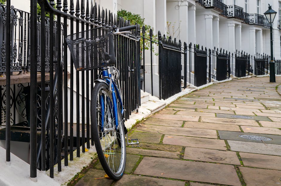 A close-up view of a residential sidewalk outside a white building featuring a black iron fence, with a blue bicycle parked against the fence. The sidewalk is paved with large rectangular stones, with