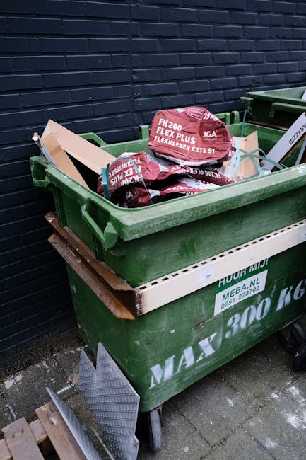 A set of four waste bins arranged outdoors on a raised platform, each with a different color lid and label for recycling categories. The blue bin on the left, designated for paper, features a recyclin