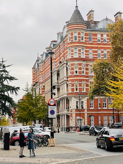A street scene in West Kensington featuring a large red-brick Victorian-style building with white architectural details, multiple bay windows, and a conical turret. The street is lined with trees showing autumn foliage, and there are parked cars along the curb. In the foreground, two people are engaged in conversation beside parked bicycles and a pedestrian crossing sign displaying a 20 mph speed limit. The scene is captured during daytime with overcast skies. While the image focuses on the exterior architecture and street activity, professional cleaning services like those offered by Cleaners West Kensington can help maintain the cleanliness of buildings and public areas in residential and commercial settings in North End Road flats, West Kensington.