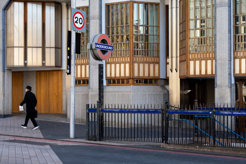 Photograph of the exterior of a modern building in South Kensington, featuring large glass windows framed by wooden panels, with a visible underground station entrance sign nearby. The sidewalk is clean and paved with rectangular tiles, and a man dressed in dark clothing and a hat is walking past holding a paper. A black metal fence runs along the edge of the pavement, separating the pedestrian walkway from the street. The scene is well-lit with natural daylight, highlighting the pristine condition of the building’s surfaces and the tidy street environment, reflecting professional cleaning and maintenance practices as provided by Cleaners West Kensington.