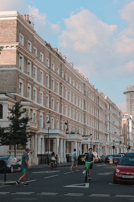 A street scene in West Kensington featuring a large red-brick Victorian-style building with white architectural details, multiple bay windows, and a conical turret. The street is lined with trees showing autumn foliage, and there are parked cars along the curb. In the foreground, two people are engaged in conversation beside parked bicycles and a pedestrian crossing sign displaying a 20 mph speed limit. The scene is captured during daytime with overcast skies. While the image focuses on the exterior architecture and street activity, professional cleaning services like those offered by Cleaners West Kensington can help maintain the cleanliness of buildings and public areas in residential and commercial settings in North End Road flats, West Kensington.